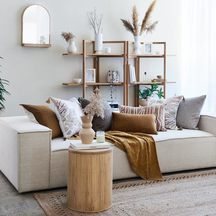 Living room image with wooden coffee table, yellow throw on white couch with a selection of earthen coloured cushions and dried flowers in the background