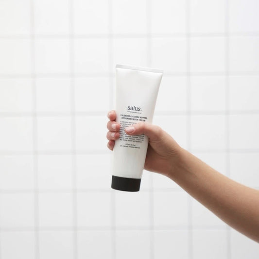 Person holding a bottle of Salus body cream in front of a white wall